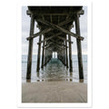 Timber pier viewed from beneath, with crossed wooden beams receding over gentle sea waves and wet sand under a grey sky.