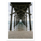 Timber pier viewed from beneath, with crossed wooden beams receding over gentle sea waves and wet sand under a grey sky.