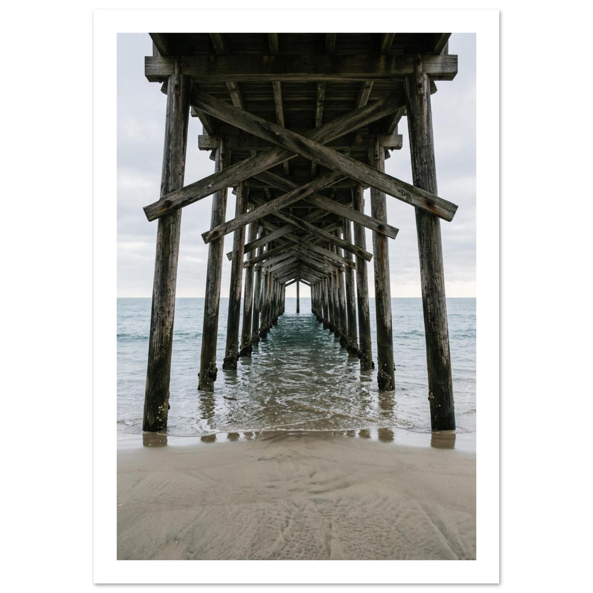 Timber pier viewed from beneath, with crossed wooden beams receding over gentle sea waves and wet sand under a grey sky.