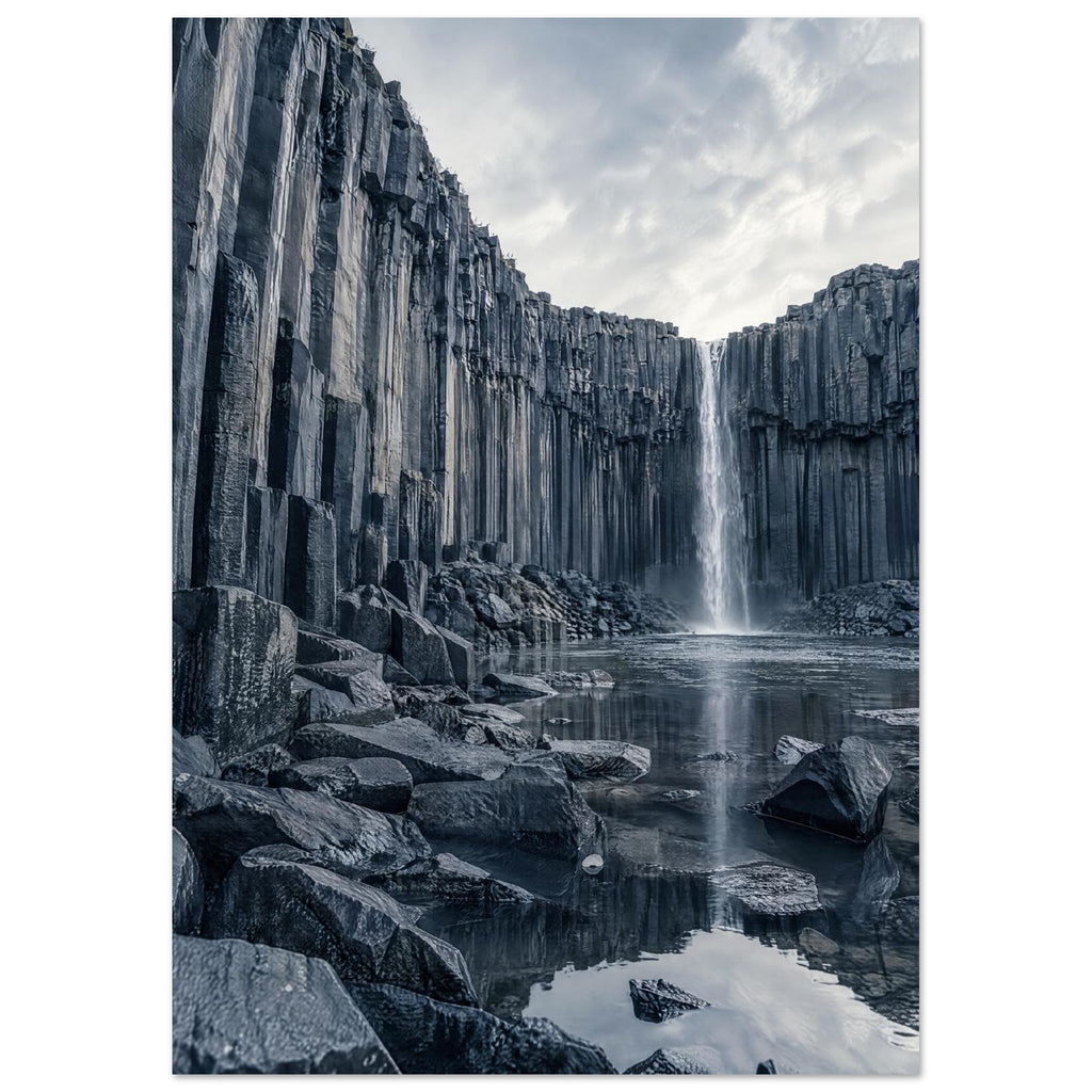 Waterfall cascading between tall basalt columns into a reflective pool, with dark foreground rocks and a cloudy sky.