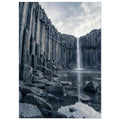 Waterfall cascading between tall basalt columns into a reflective pool, with dark foreground rocks and a cloudy sky.
