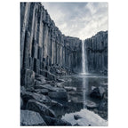 Waterfall cascading between tall basalt columns into a reflective pool, with dark foreground rocks and a cloudy sky.
