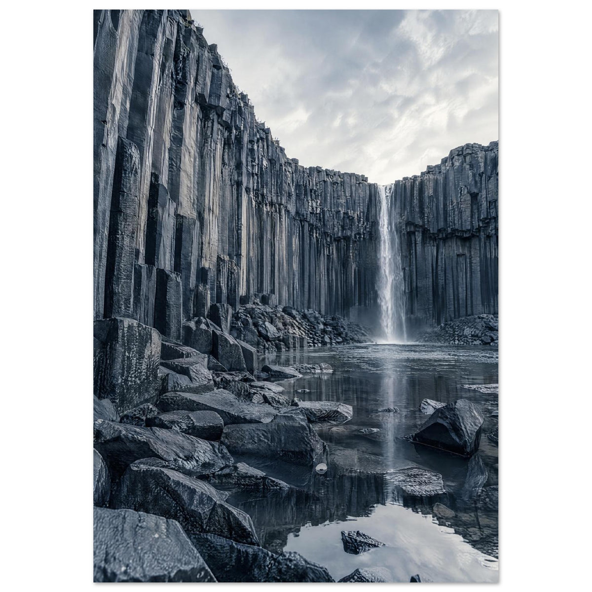Waterfall cascading between tall basalt columns into a reflective pool, with dark foreground rocks and a cloudy sky.