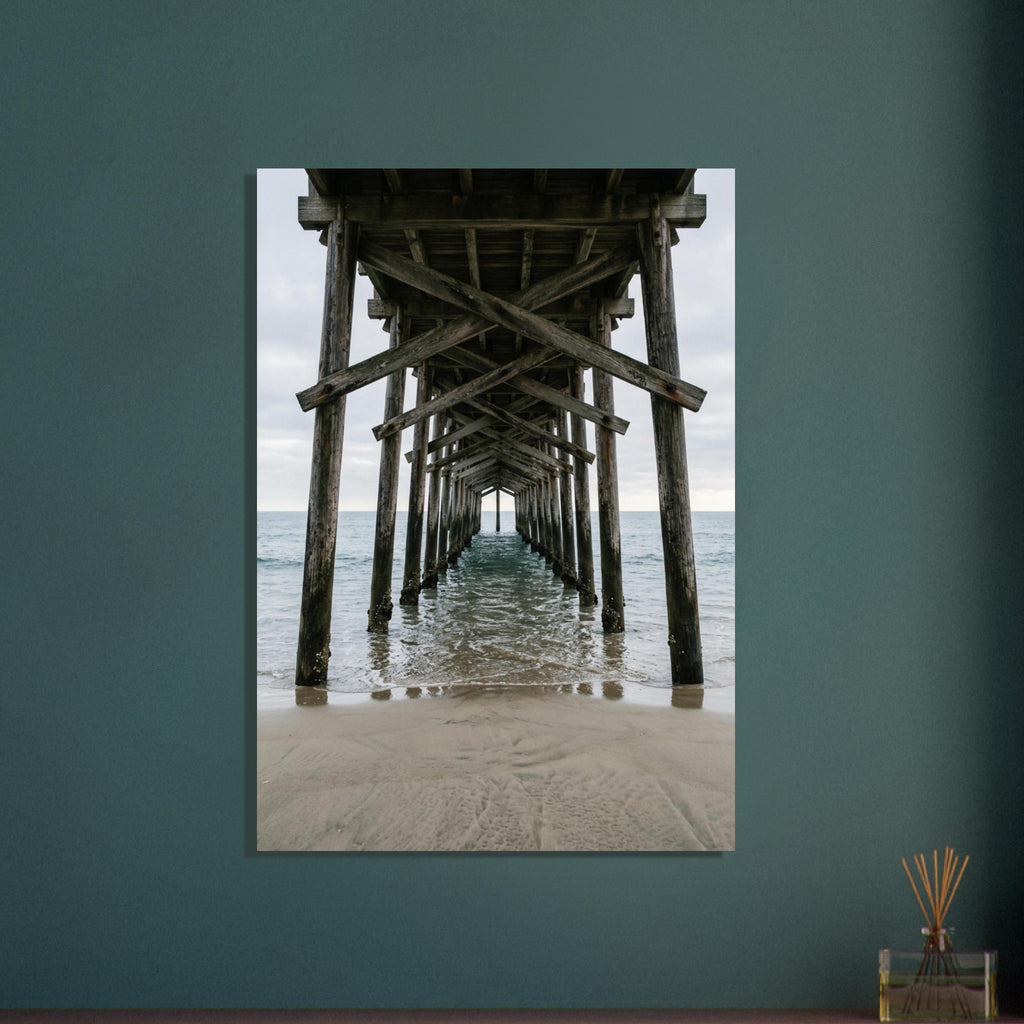 Timber pier viewed from beneath, with crossed wooden beams receding over gentle sea waves and wet sand under a grey sky.