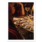 Oysters on ice with lemon wedges, sparkling wine and a glowing lamp beside a cropped figure in a burgundy dress.