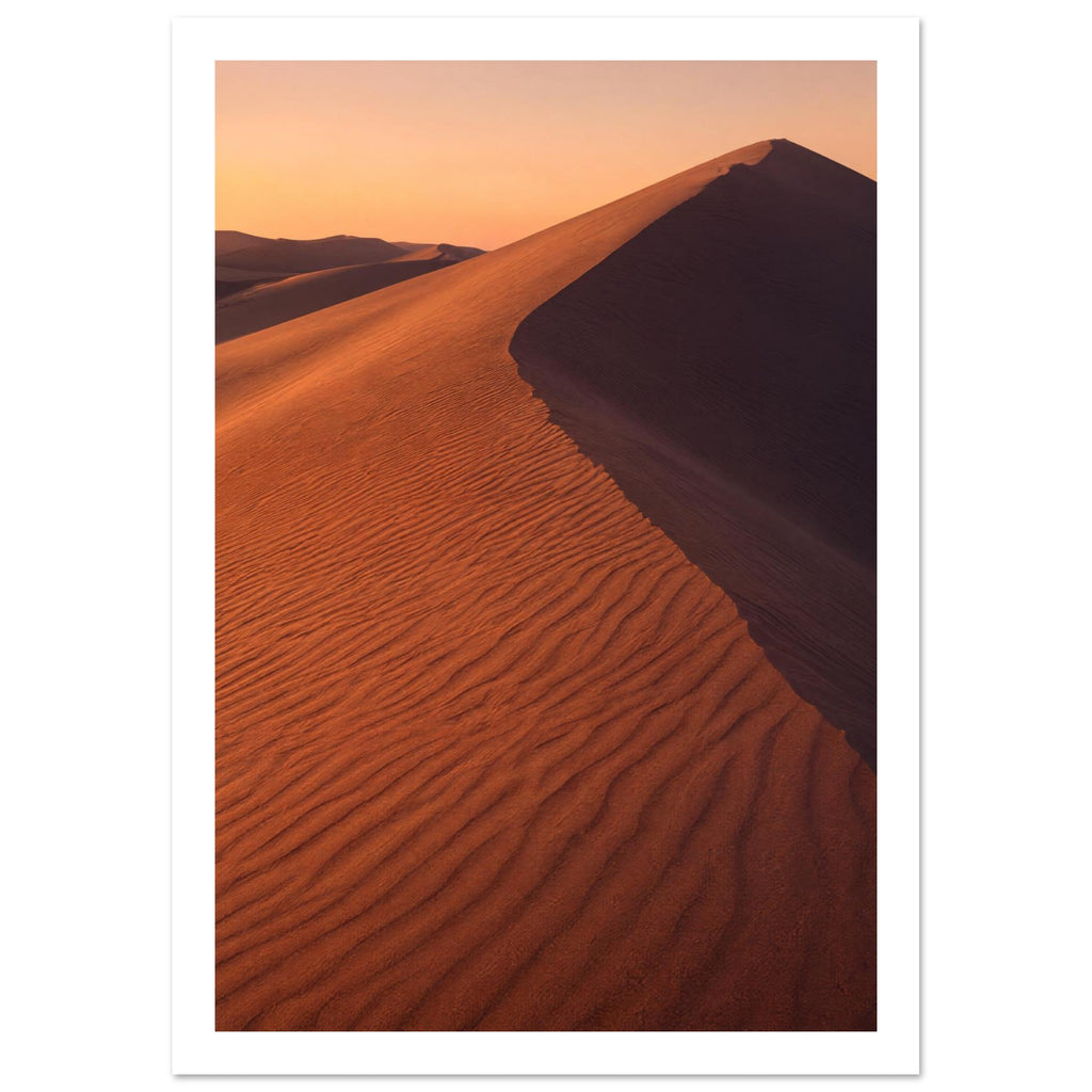Desert sand dune with rippled orange slopes, a sweeping ridge and deep brown shadow beneath a peach sky.