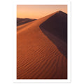 Desert sand dune with rippled orange slopes, a sweeping ridge and deep brown shadow beneath a peach sky.