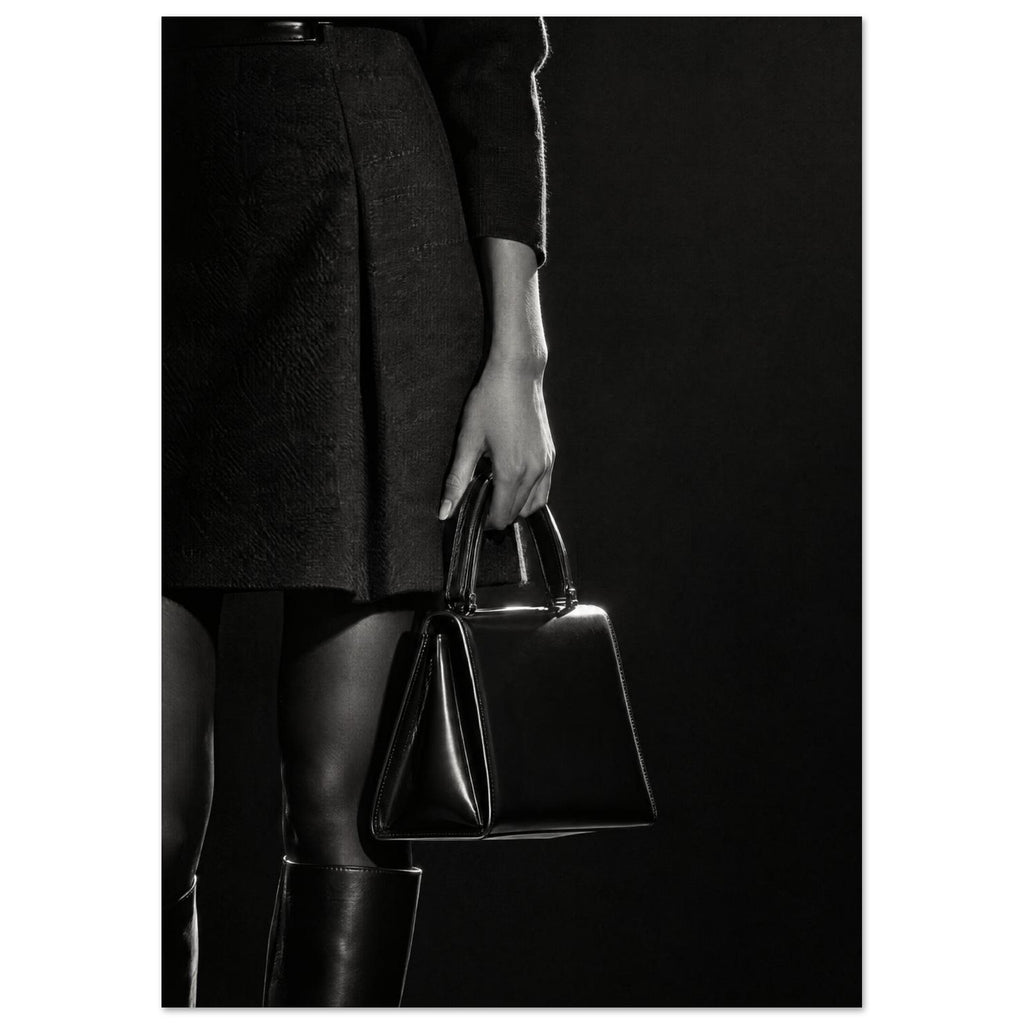 Cropped black and white photograph of a figure in a dark skirt and boots holding a glossy structured handbag.