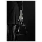 Cropped black and white photograph of a figure in a dark skirt and boots holding a glossy structured handbag.