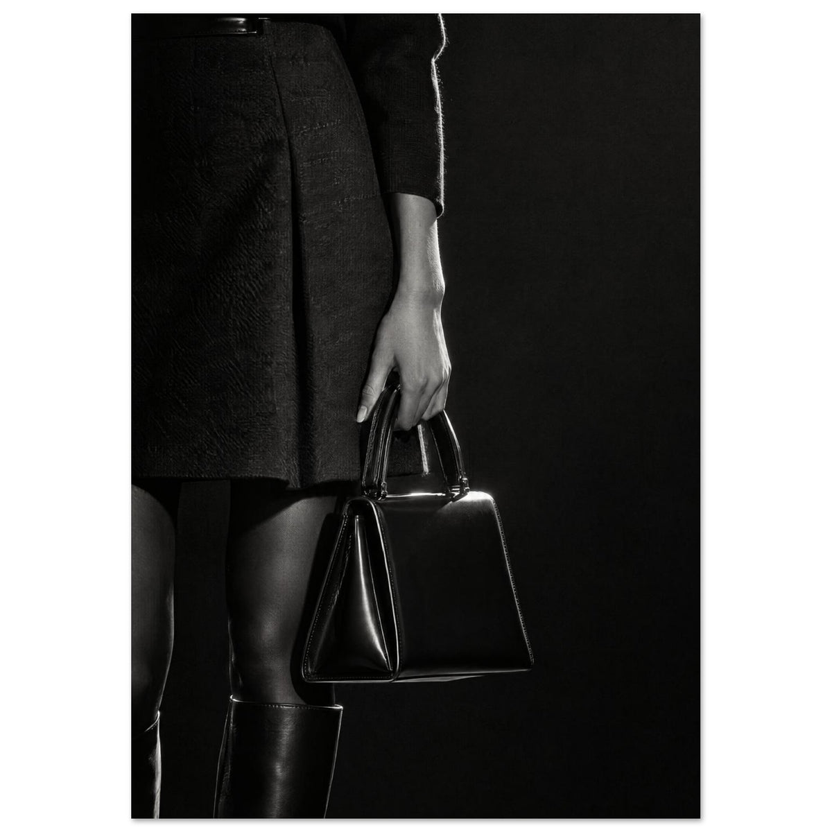Cropped black and white photograph of a figure in a dark skirt and boots holding a glossy structured handbag.