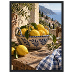 Lemons in a blue-and-white bowl on a wooden table, with olive branches, checked cloth and a coastal village beyond.