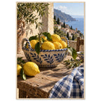 Lemons in a blue-and-white bowl on a wooden table, with olive branches, checked cloth and a coastal village beyond.