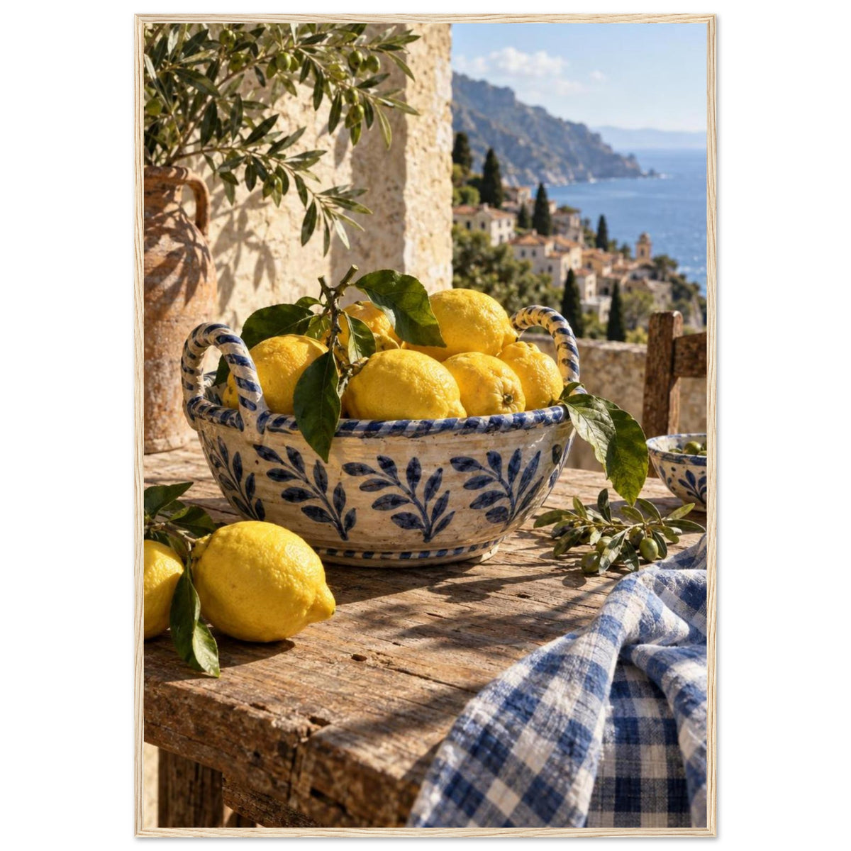 Lemons in a blue-and-white bowl on a wooden table, with olive branches, checked cloth and a coastal village beyond.