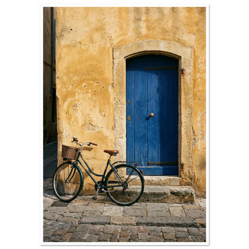 Green bicycle with a woven basket beside a blue wooden door in a golden plaster wall, above worn stone paving.