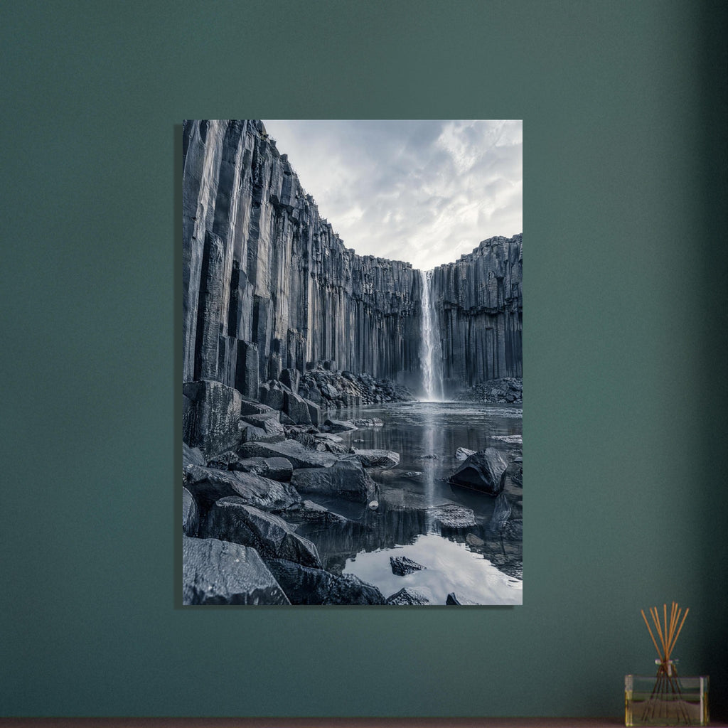 Waterfall cascading between tall basalt columns into a reflective pool, with dark foreground rocks and a cloudy sky.