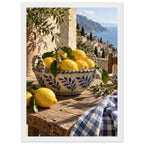 Lemons in a blue-and-white bowl on a wooden table, with olive branches, checked cloth and a coastal village beyond.