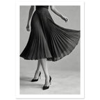 Cropped figure in a flowing black pleated skirt and pointed high heels, with crossed legs against a grey background.