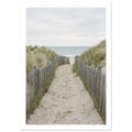 Sandy path with footprints between wooden fences and grassy dunes, leading towards a pale blue sea beneath a cloudy sky.