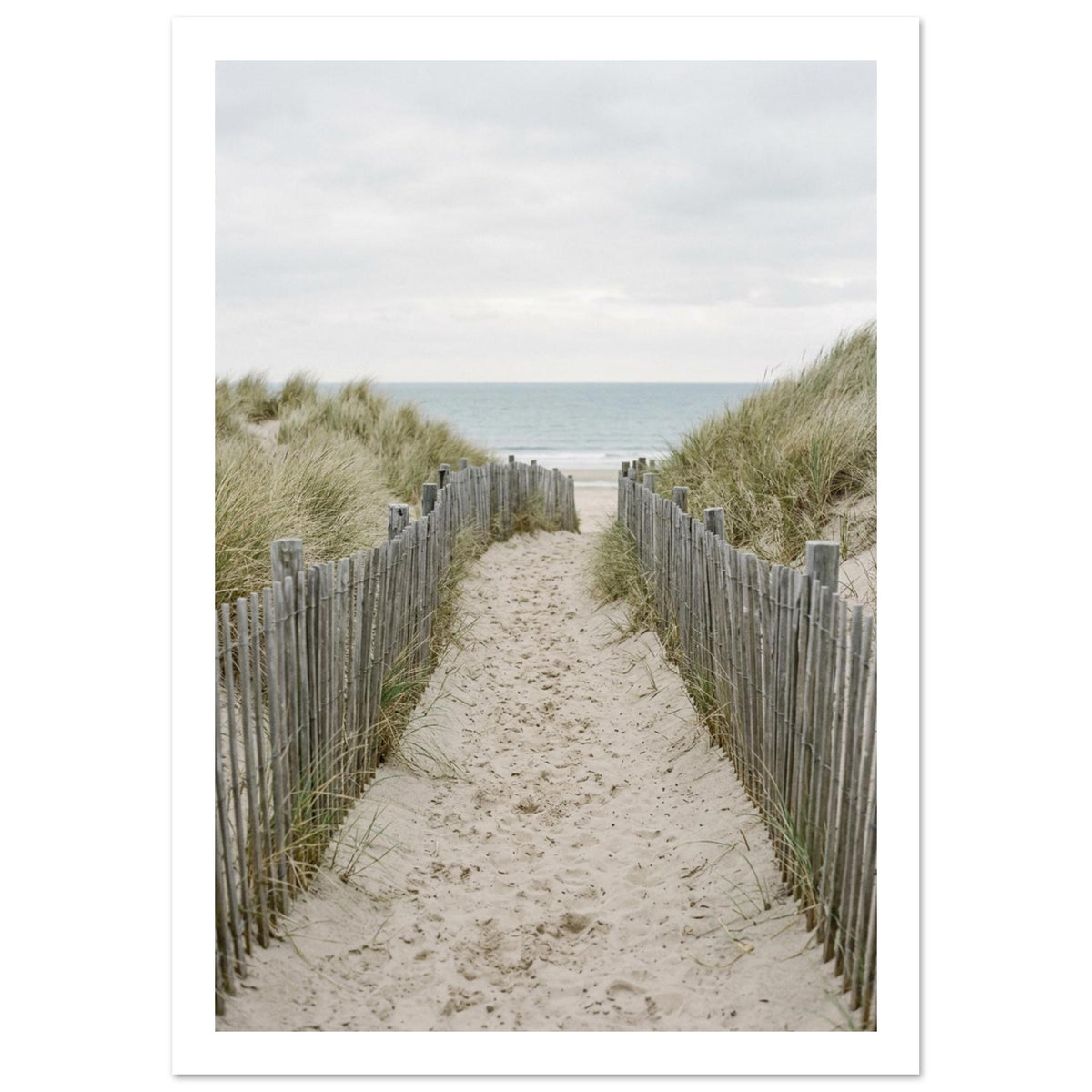 Sandy path with footprints between wooden fences and grassy dunes, leading towards a pale blue sea beneath a cloudy sky.