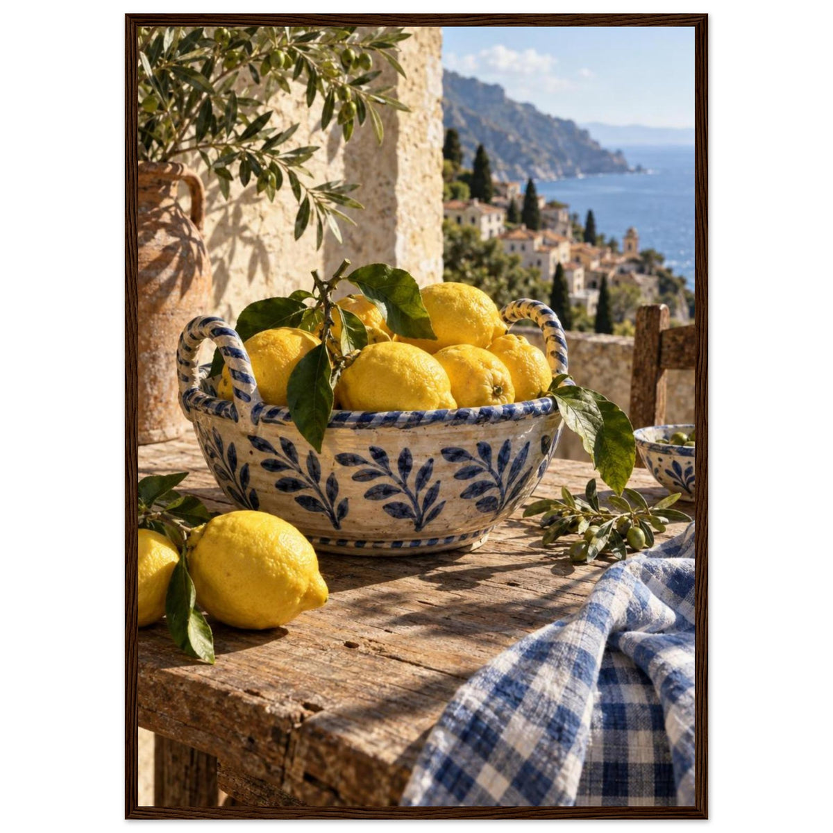 Lemons in a blue-and-white bowl on a wooden table, with olive branches, checked cloth and a coastal village beyond.