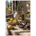 Lemons, olives, figs and a bottle of oil on a stone table beside a ceramic jug of leafy branches and a sunlit window.