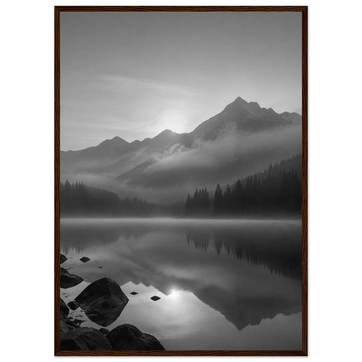 Mountain peaks and conifer forests reflected in a misty lake, with rocks in the foreground, in black and white.