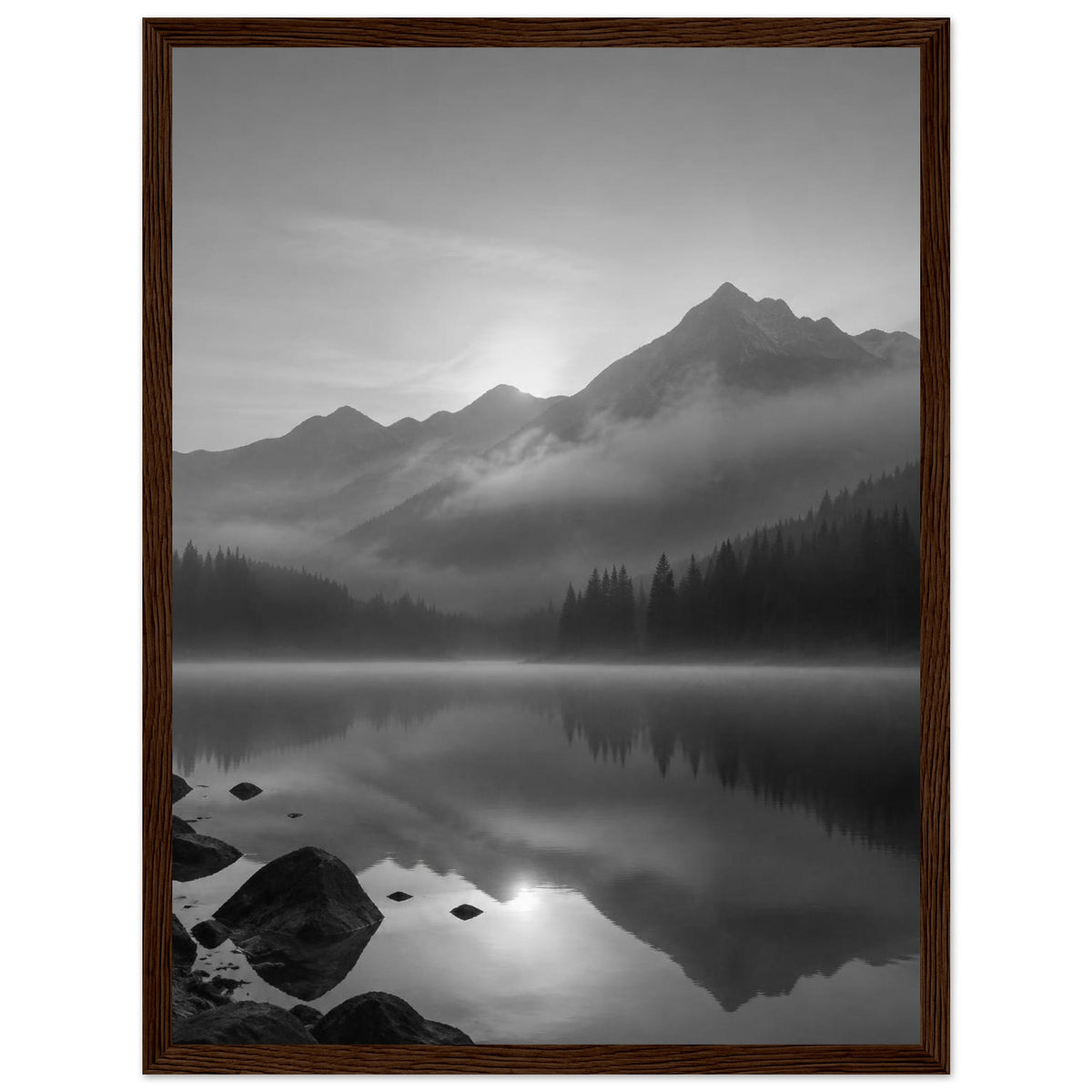 Mountain peaks and conifer forests reflected in a misty lake, with rocks in the foreground, in black and white.