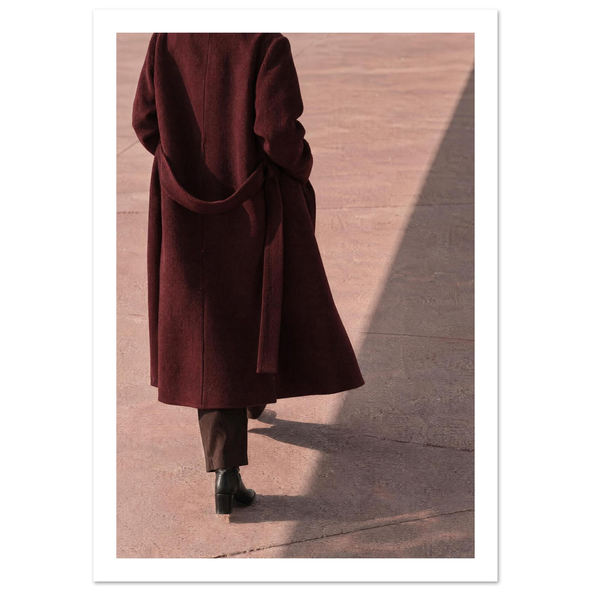 Figure seen from behind in a long burgundy coat, brown trousers and black boots on pink pavement with diagonal shadows.