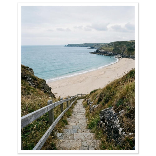 Stone steps with a wooden handrail descend through coastal grasses to a sandy beach, blue sea and rocky headlands.