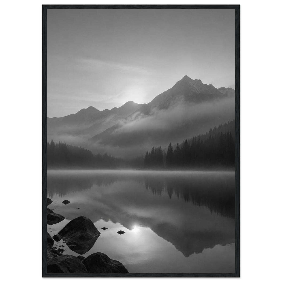 Mountain peaks and conifer forests reflected in a misty lake, with rocks in the foreground, in black and white.