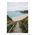 Stone steps with a wooden handrail descend through coastal grasses to a sandy beach, blue sea and rocky headlands.